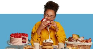 mulher diante de uma mesa repleta de doces comendo uma torta e sentindo fome ou gula