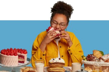 mulher diante de uma mesa repleta de doces comendo uma torta e sentindo fome ou gula